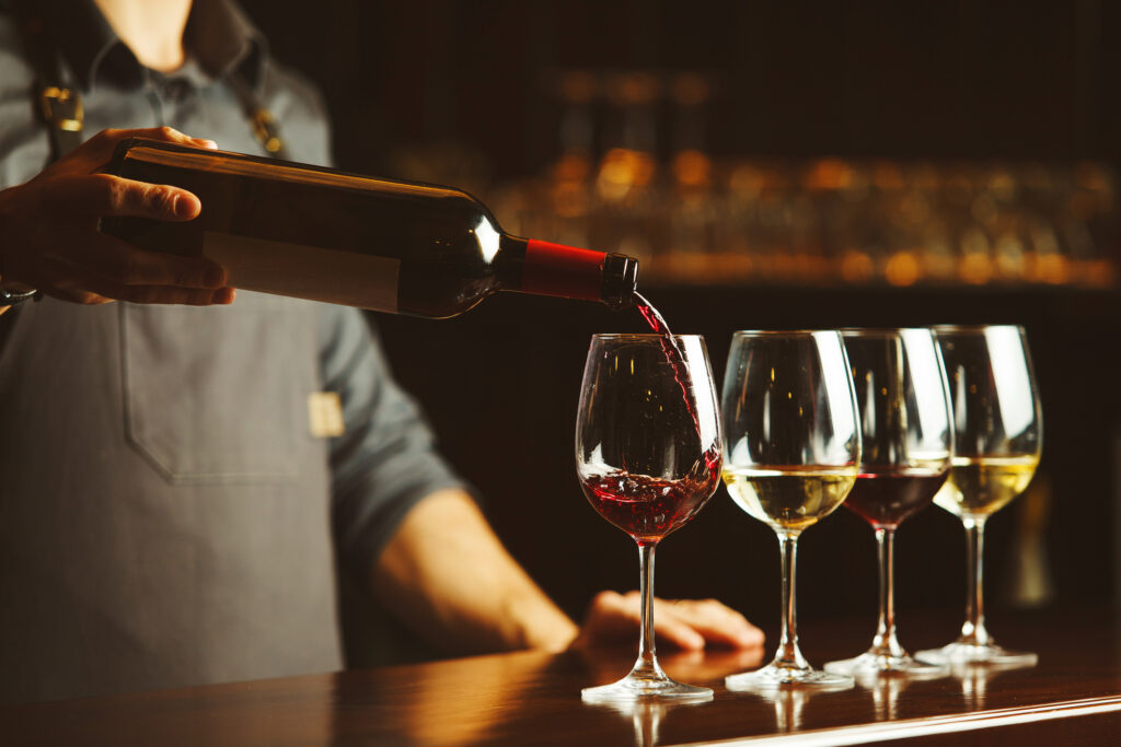 Bartender in apron pours sweet red wine from bottle in capacious glasses on wooden bar counter. Delicious alcohol drink in transparent glassware with nice scent.